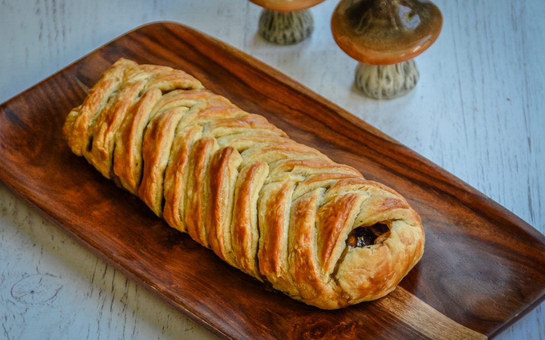 Golden braided mushroom strudel on a wooden platter beside two ceramic mushroom ornaments.