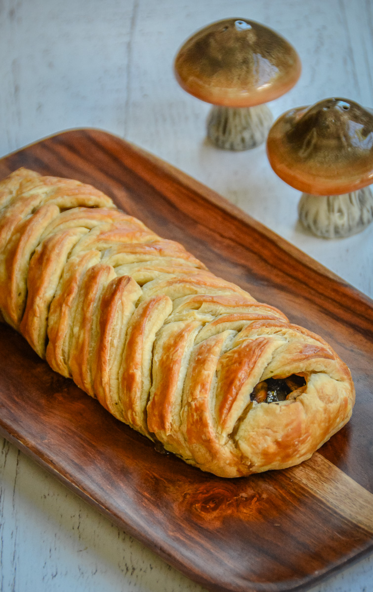 Close view of mushroom strudel showing overlapping strips of golden puff pastry on a wooden platter.