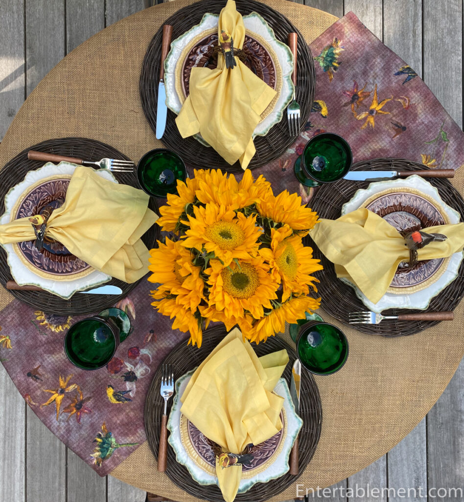 Overhead view of a round late-summer table with sunflowers, yellow napkins, green goblets and layered place settings.