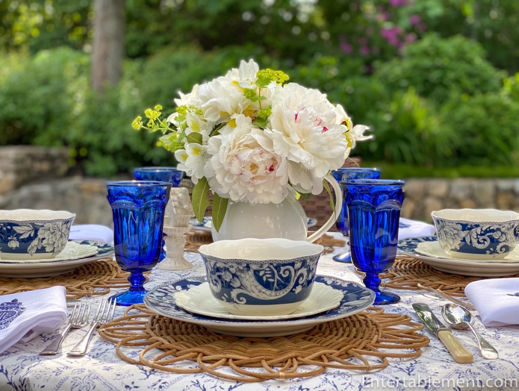 Adelaide in Navy dinner plate by 222 Fifth featuring white floral and bird motif on a deep indigo background, placed on a rattan charger.