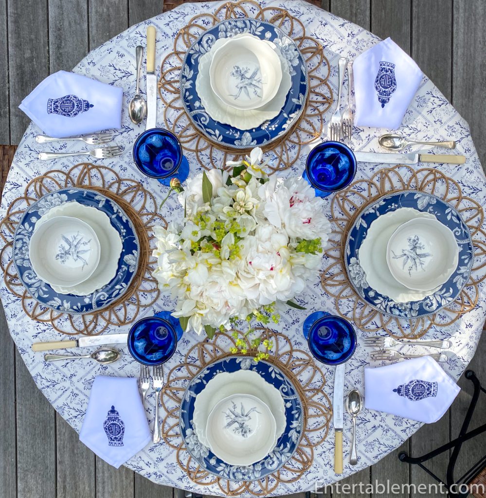 Overhead view of blue and white summer tablescape with Adelaide dinnerware, cobalt goblets, peony centerpiece, and rattan placemats.