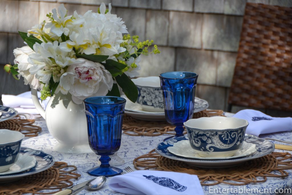 Adelaide in Navy dinner plate by 222 Fifth featuring white floral and bird motif on a deep indigo background, placed on a rattan charger.