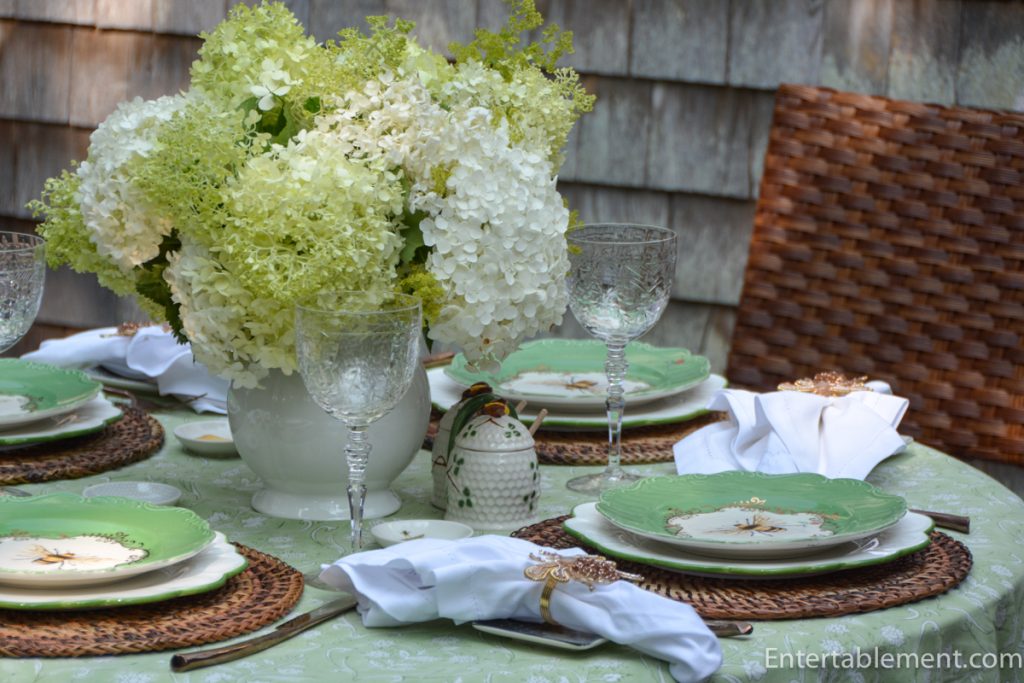 Fresh Limelight hydrangeas arranged in a white pitcher surrounded by Anthropologie bee plates, crystal goblets and woven chargers for outdoor summer dining. 