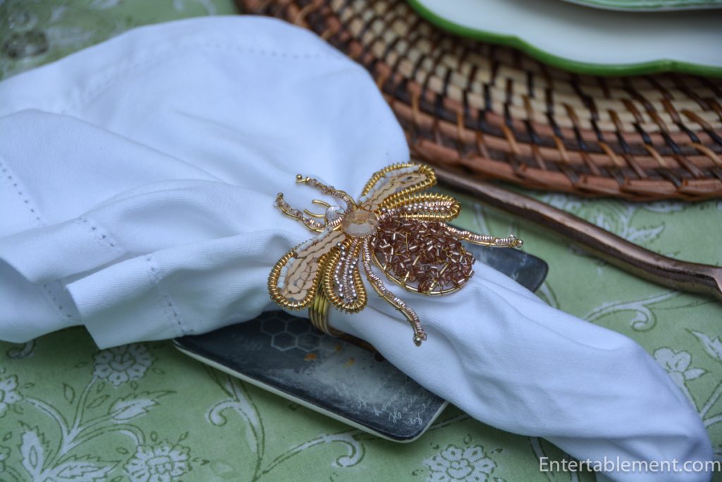 Gold beaded bee napkin ring on a crisp white linen napkin with antique gold flatware and woven charger beneath.