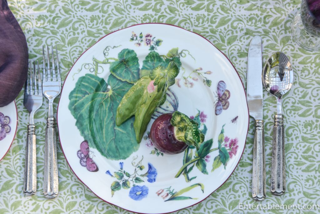 Chelsea porcelain dinner plate with green leaves, flowers and butterflies on a pale green patterned tablecloth, set with purple linen, green glass candlesticks and ceramic aubergines.