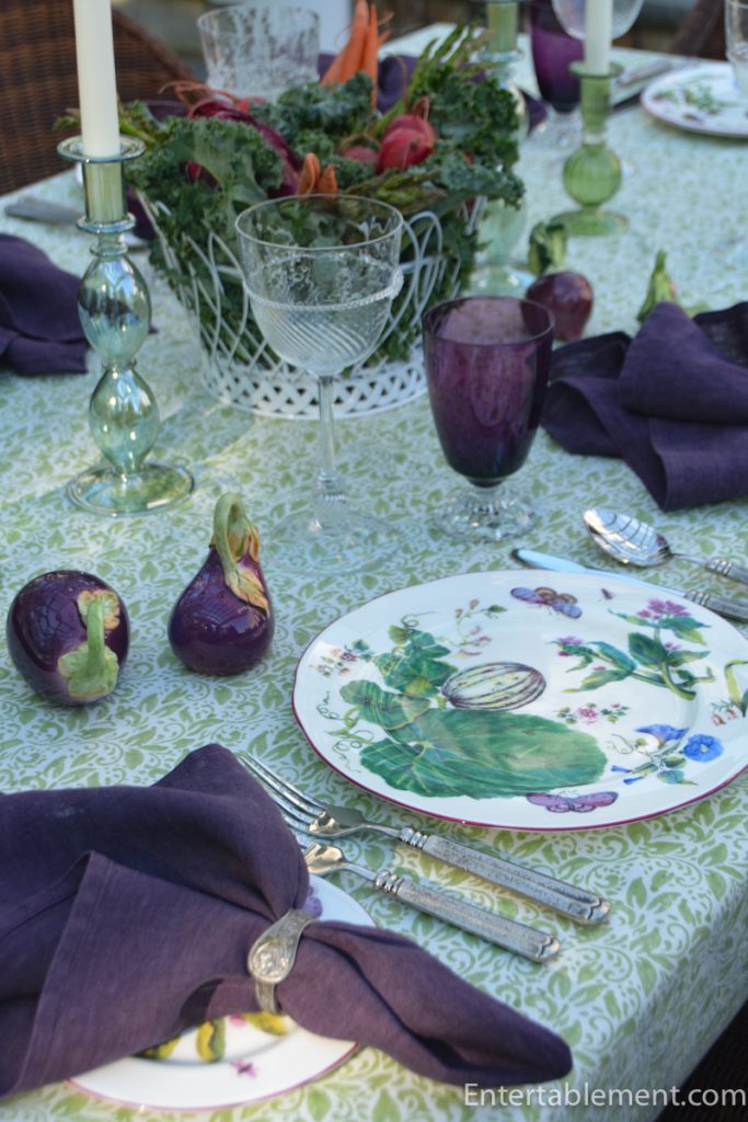 Purple footed glass beside ceramic aubergine ornaments and a green glass candlestick on a pale green patterned outdoor tablecloth.