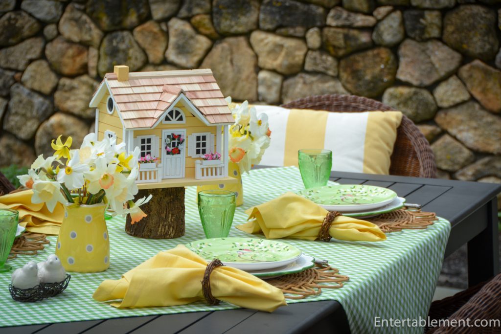 Outdoor spring tablescape with birdhouse centerpiece, daffodils, green glassware, and yellow napkins