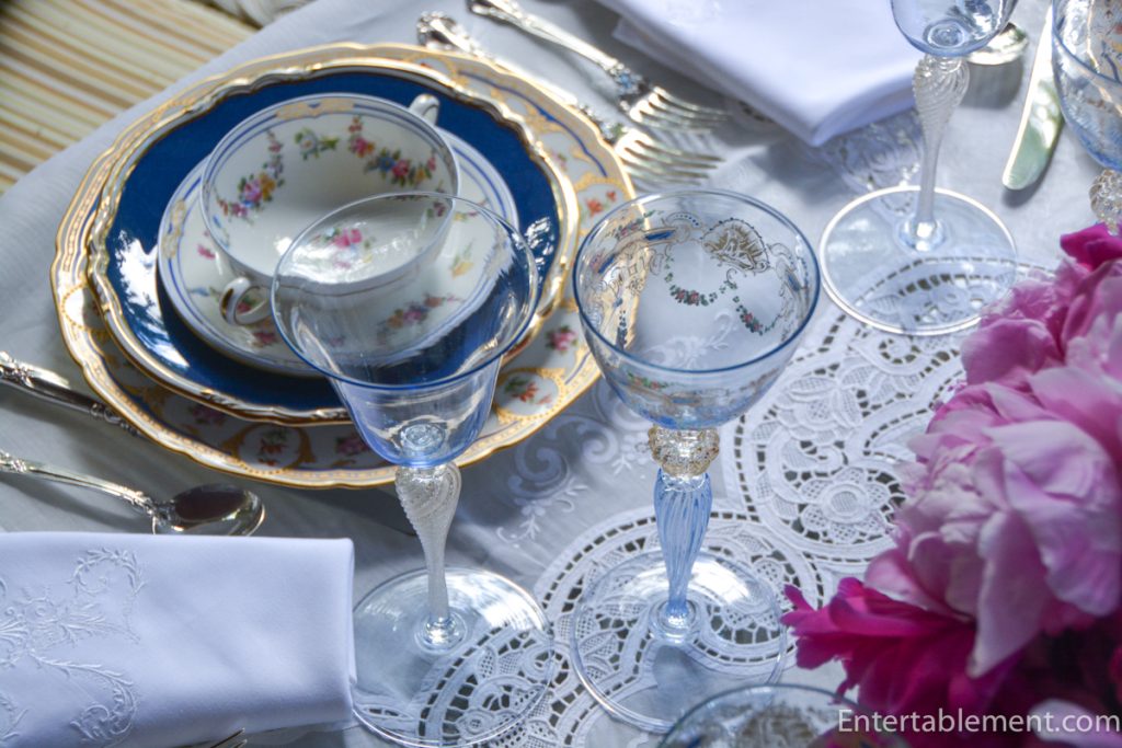 Antique Venetian goblet with gold detailing beside embroidered white napkin and pink peonies.
