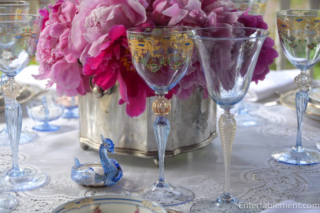 Antique Venetian goblet with gold detailing beside embroidered white napkin and pink peonies.
