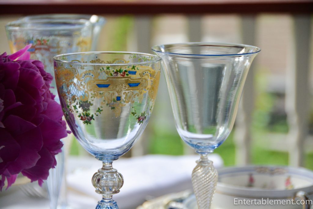Antique Venetian goblet with gold detailing beside embroidered white napkin and pink peonies.
