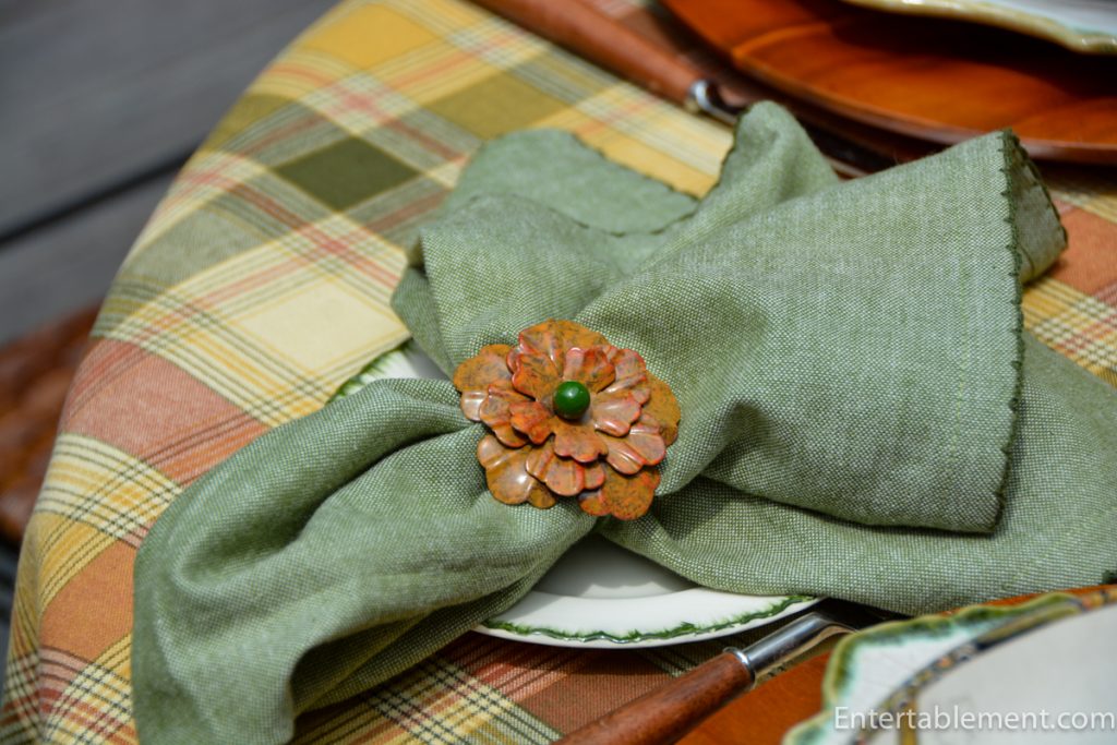 Green linen napkin with floral copper ring beside amber-stemmed goblet on fall plaid tablecloth.