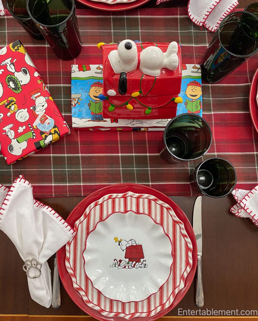 Peanuts Christmas plate showing Snoopy atop a red doghouse surrounded by peppermint candies, paired with red striped and solid red chargers.