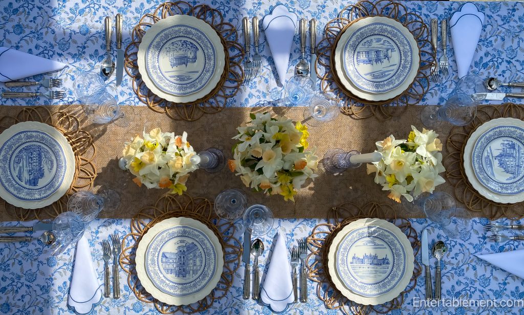 A spring tablescape featuring Gien Château series plates, blue and white floral linens, rattan chargers, etched glassware, and yellow daffodils, set outdoors on a breezy afternoon.