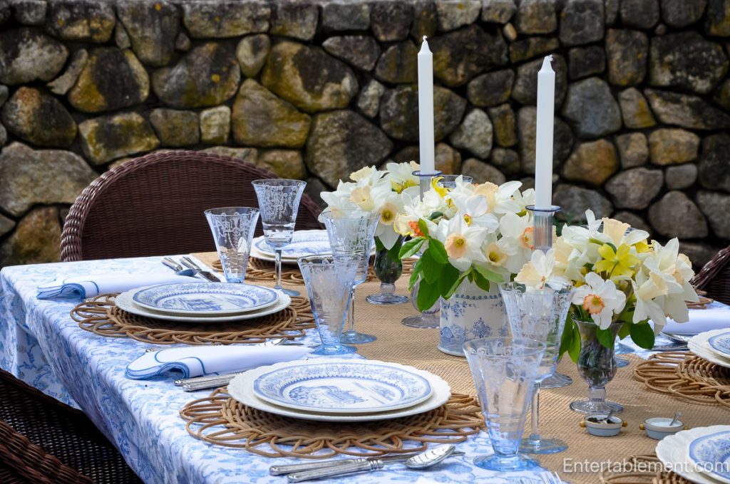 A spring tablescape featuring Gien Château series plates, blue and white floral linens, rattan chargers, etched glassware, and yellow daffodils, set outdoors on a breezy afternoon.