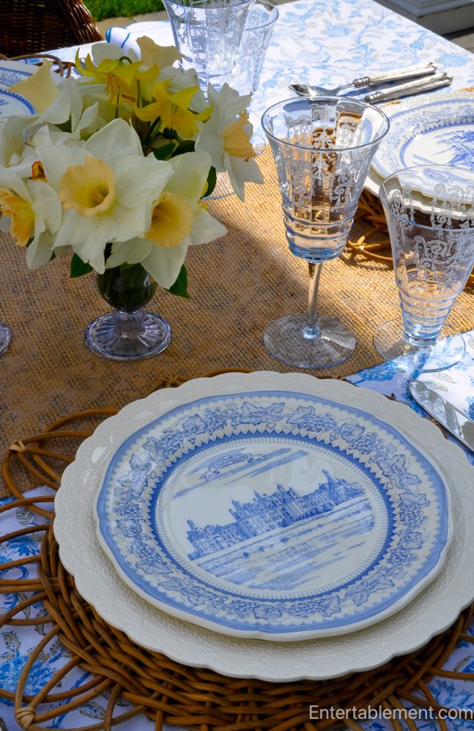 A spring tablescape featuring Gien Château series plates, blue and white floral linens, rattan chargers, etched glassware, and yellow daffodils, set outdoors on a breezy afternoon.