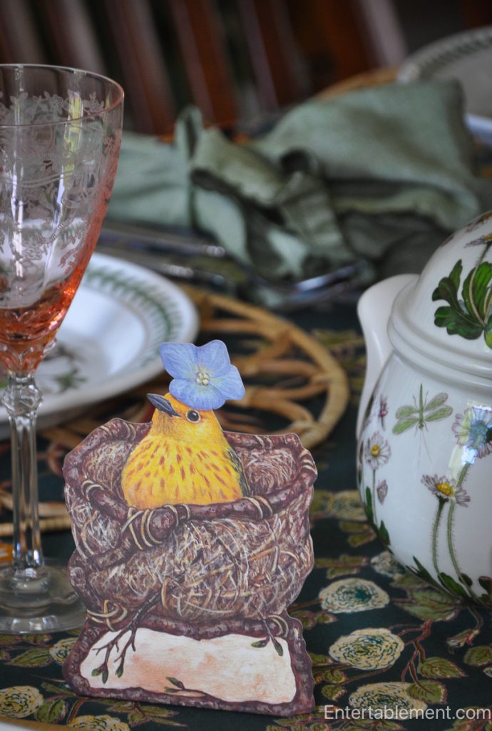 Paper place card of a yellow bird in a nest, wearing a blue flower, next to pink etched glassware.