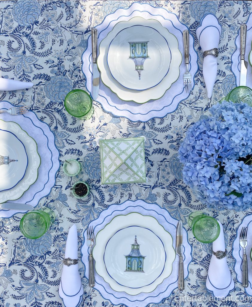 Tablescape styled with Mrs. Alice blue pagoda plates, paired with scalloped white placemats by Rebecca Udall and vintage pink Cherry Blossom glassware by Jeanette. The centerpiece features lush blue hydrangeas in a white vessel, flanked by classic blue-and-white chinoiserie lanterns from The Enchanted Home. An elegant blend of whimsical and formal, set outdoors for a summer luncheon.