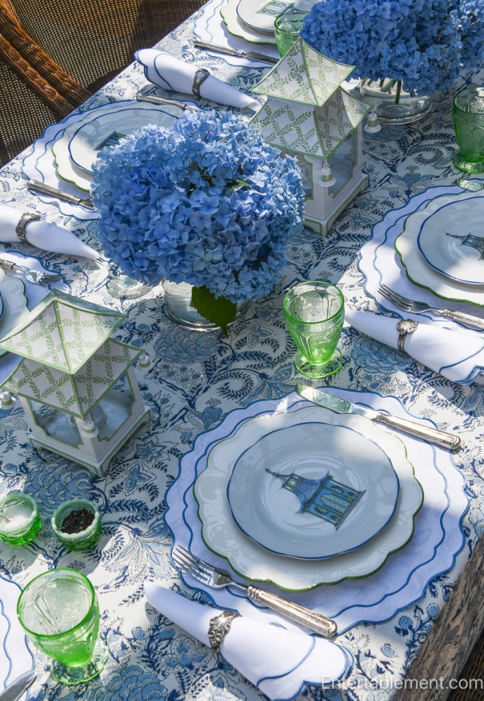 Tablescape styled with Mrs. Alice blue pagoda plates, paired with scalloped white placemats by Rebecca Udall and vintage pink Cherry Blossom glassware by Jeanette. The centerpiece features lush blue hydrangeas in a white vessel, flanked by classic blue-and-white chinoiserie lanterns from The Enchanted Home. An elegant blend of whimsical and formal, set outdoors for a summer luncheon.