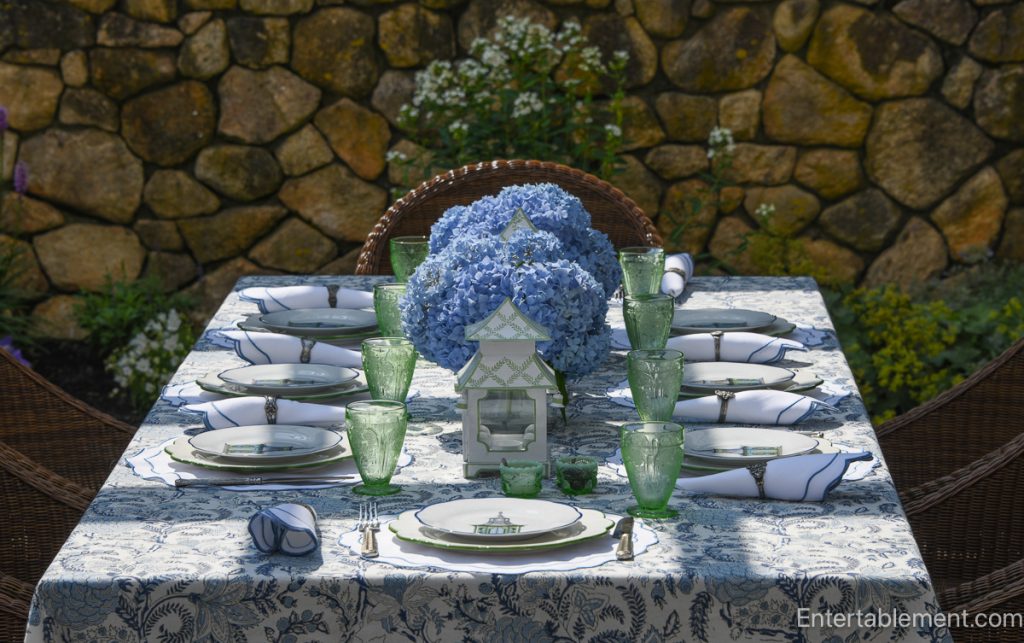 Tablescape styled with Mrs. Alice blue pagoda plates, paired with scalloped white placemats by Rebecca Udall and vintage pink Cherry Blossom glassware by Jeanette. The centerpiece features lush blue hydrangeas in a white vessel, flanked by classic blue-and-white chinoiserie lanterns from The Enchanted Home. An elegant blend of whimsical and formal, set outdoors for a summer luncheon.