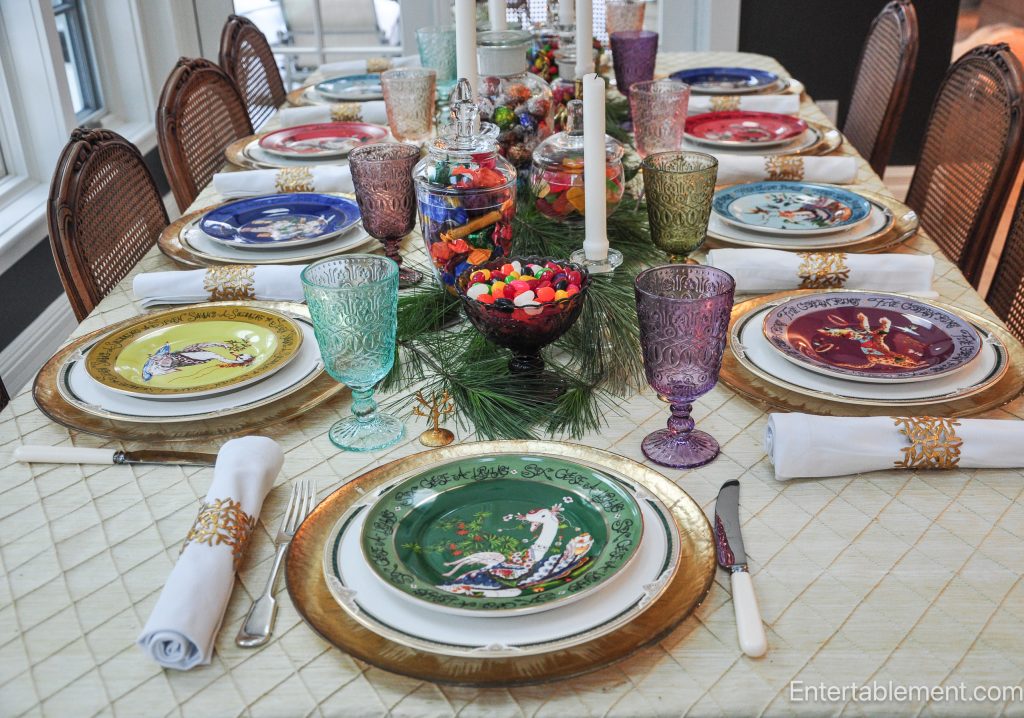 Long dining table set for Christmas with Anthropologie’s 12 Days of Christmas plates, gold chargers, multicoloured pressed-glass goblets, and a candy-filled centrepiece.