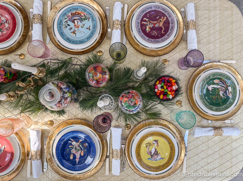 Overhead view of Christmas table featuring 12 Days of Christmas plates in assorted colours, pine greenery, candles, and jars of sweets.