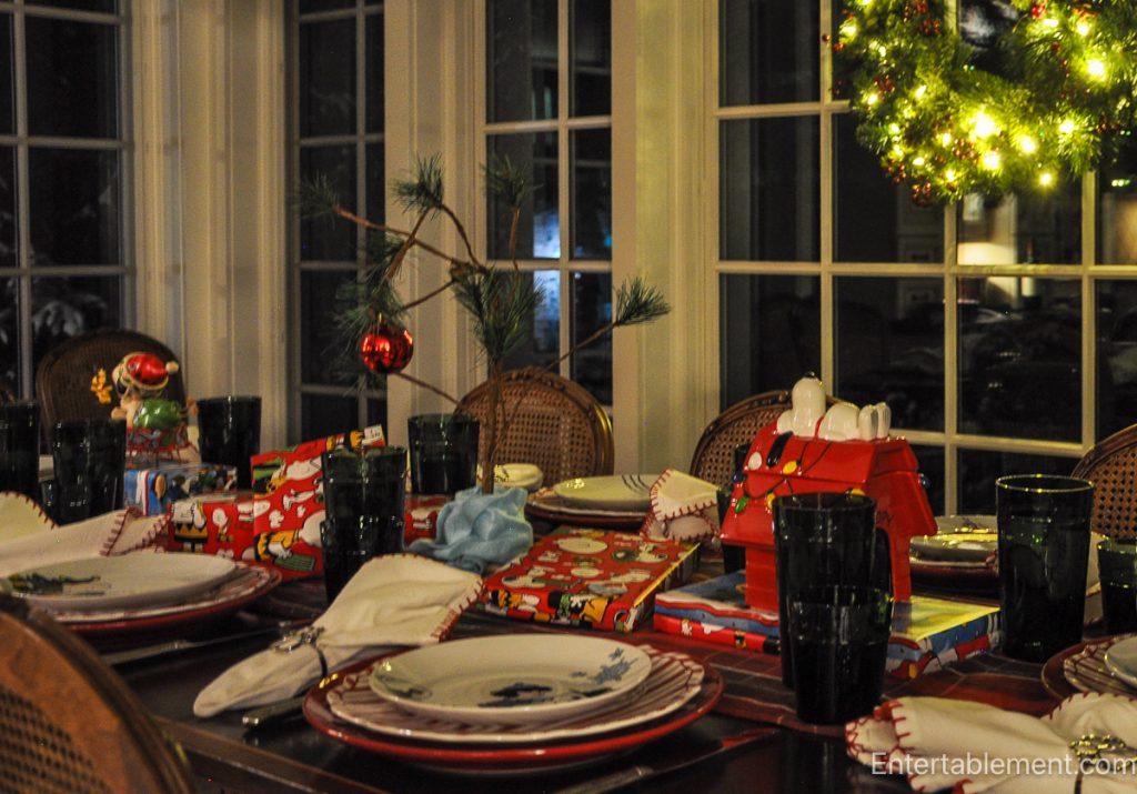 Christmas evening tablescape featuring Snoopy in a sleigh centerpiece, Peanuts-wrapped gifts, and a glowing wreath hanging in the window.
