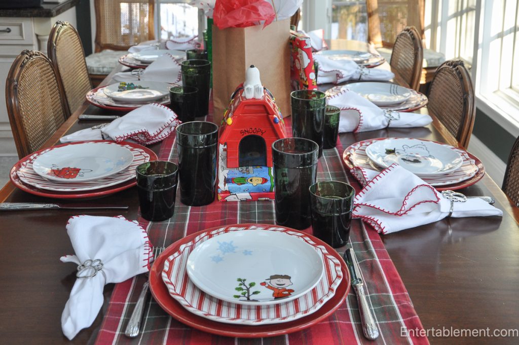 Festive Christmas table set with Snoopy doghouse cookie jar, green glassware, and Peanuts-themed dinner plates arranged around the table.