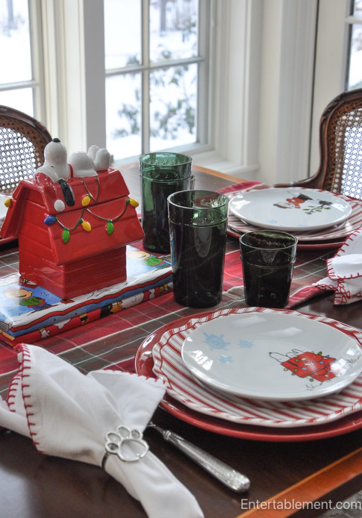 Festive Christmas table set with Snoopy doghouse cookie jar, green glassware, and Peanuts-themed dinner plates arranged around the table.