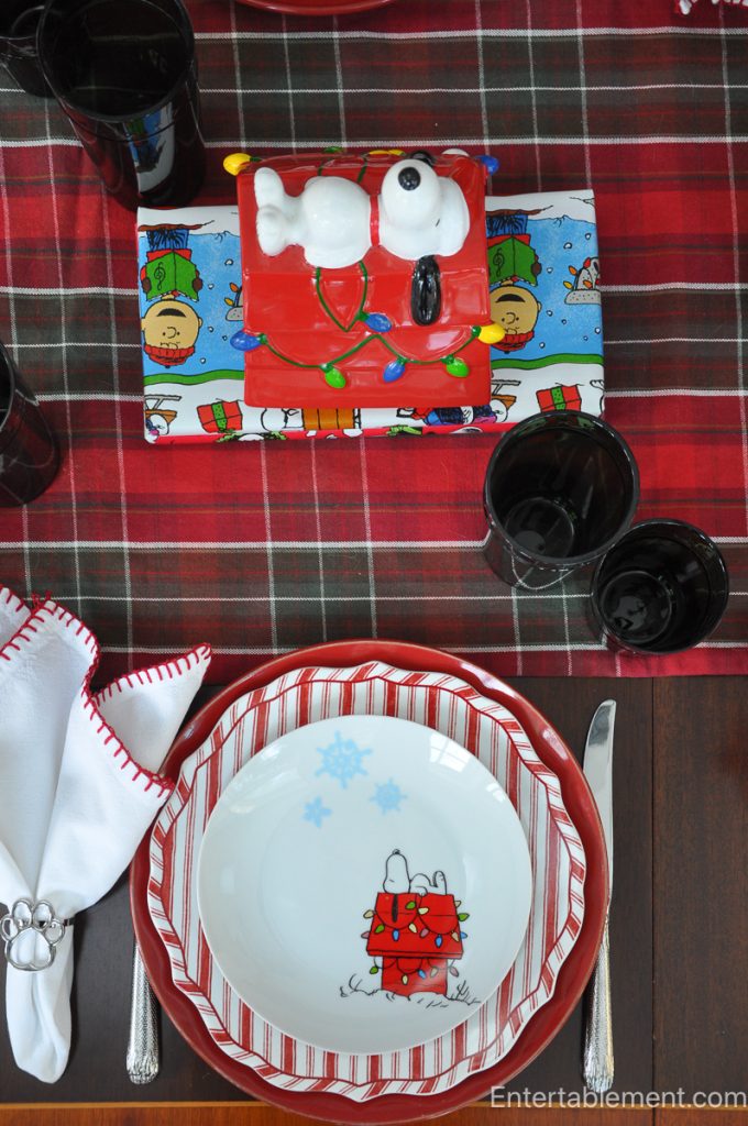Red Snoopy doghouse cookie jar surrounded by black glassware, set on a red plaid table runner with Peanuts-wrapped gifts as table accents.