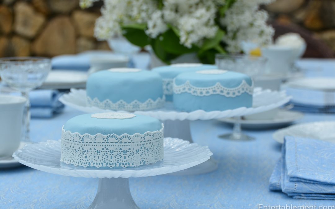 Small pale blue fondant cakes with intricate white lace detailing on a glass cake stand