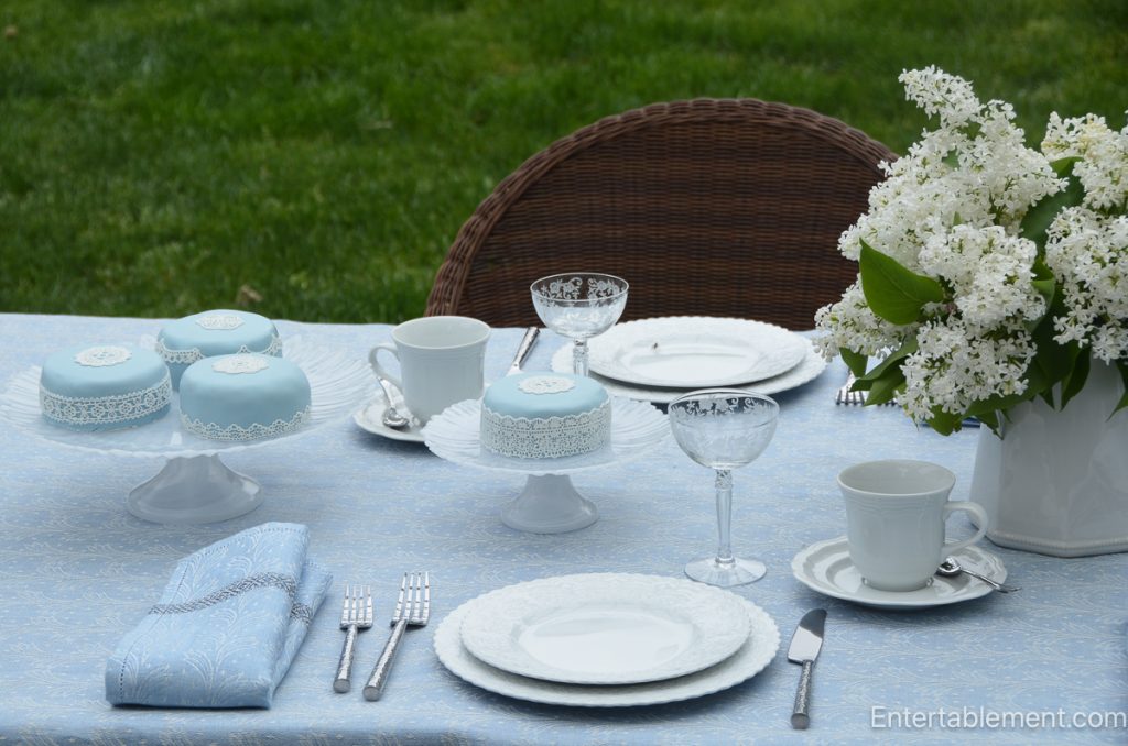 Blue and white spring tea setting with white china, lilacs, crystal glasses, and decorative cakes