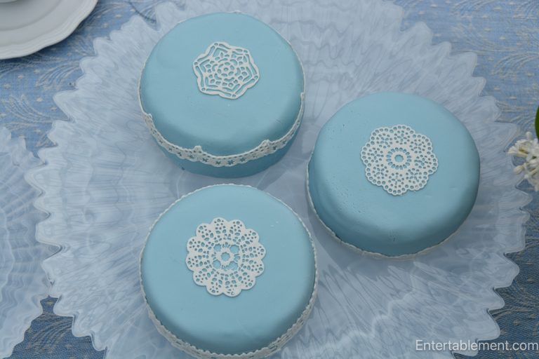 Three pale blue decorated cakes with edible lace details arranged on milk glass stands