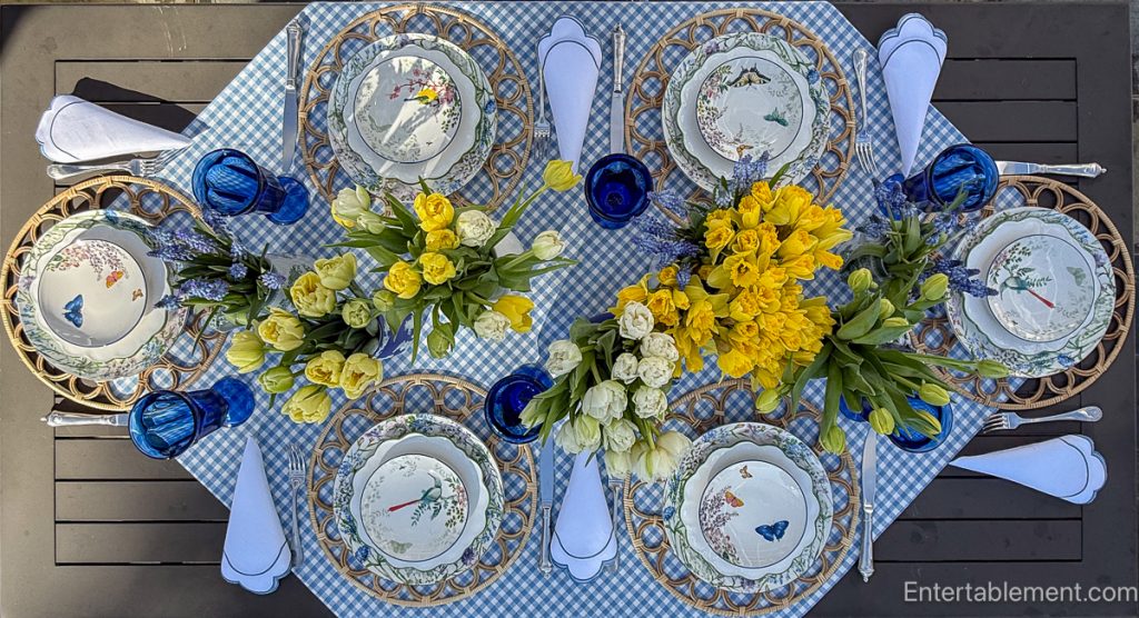 Overhead view of a spring table set outdoors with blue gingham tablecloth, woven placemats, blue goblets, and floral plates despite lingering snow