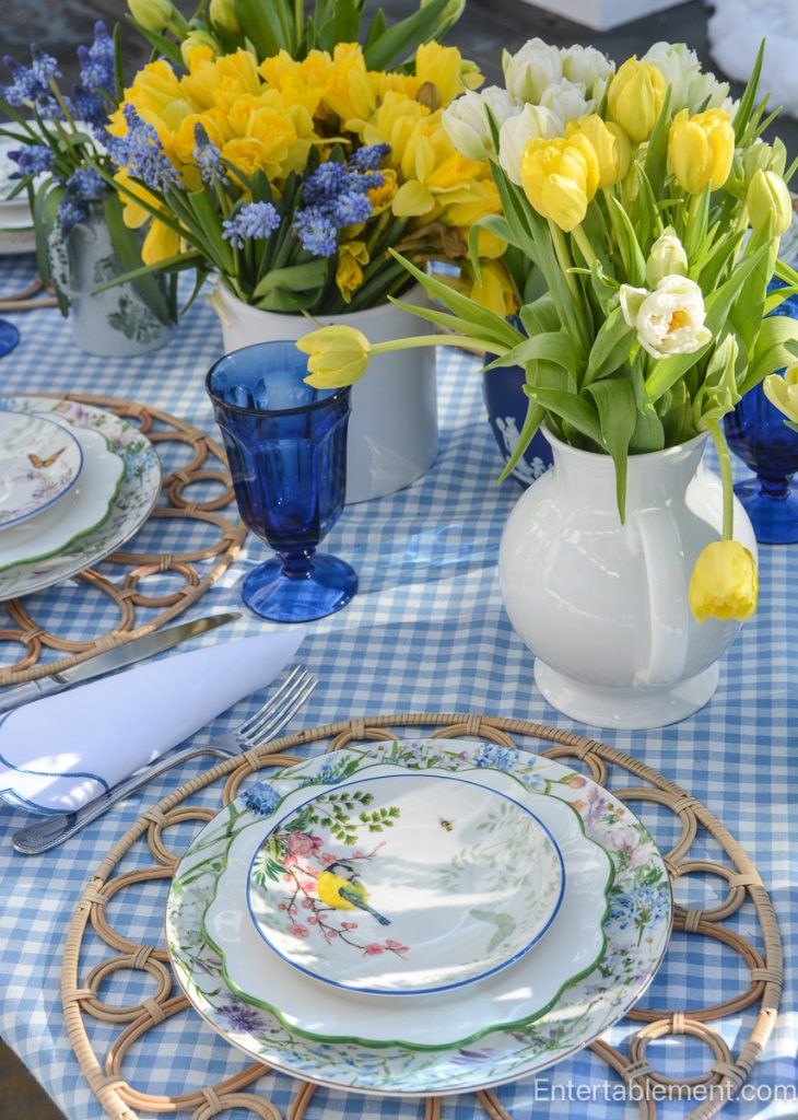 Outdoor spring table with blue and white dishes, rattan placemats, and yellow and white flowers arranged on a blue gingham cloth