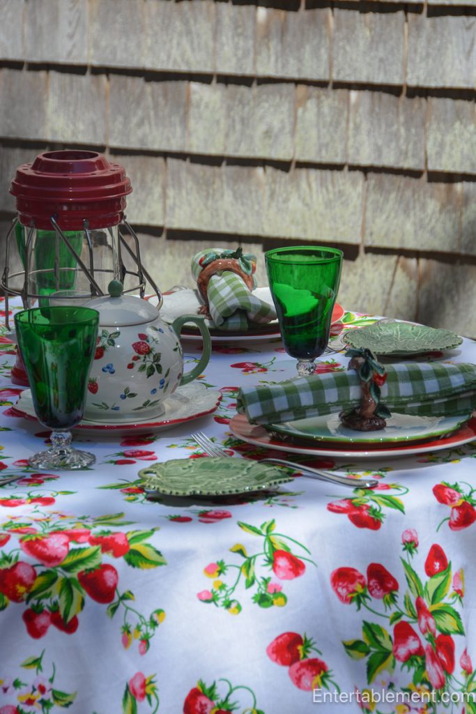 Strawberry-themed summer table with April Cornell tablecloth, green glassware, red lanterns, leafy majolica plates, and gingham napkins.