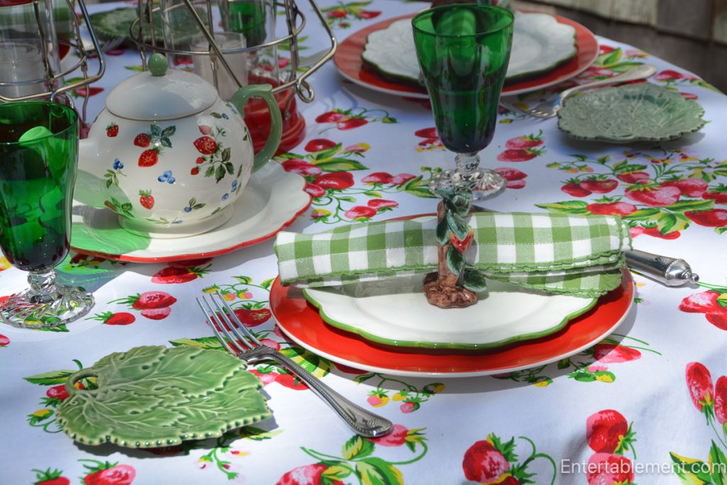 Strawberry-patterned teapot with green glassware, gingham napkins, and leafy majolica dishes on an April Cornell strawberry tablecloth.