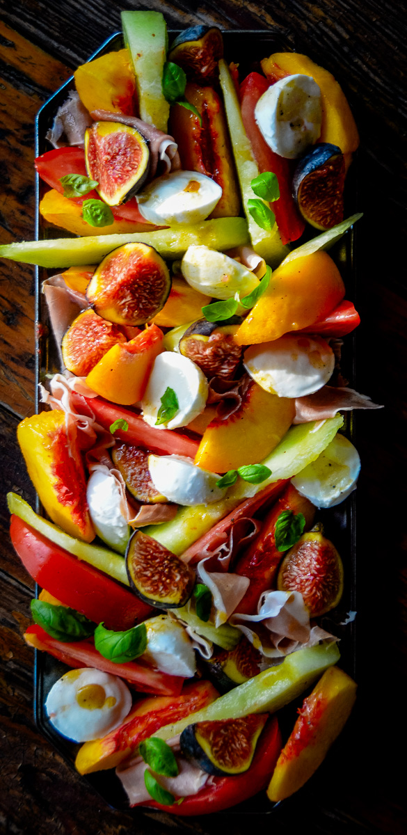 Overhead view of a long rectangular platter layered with honeydew melon, peaches, tomatoes, figs, fresh mozzarella, basil leaves, and prosciutto on a rustic wooden table.