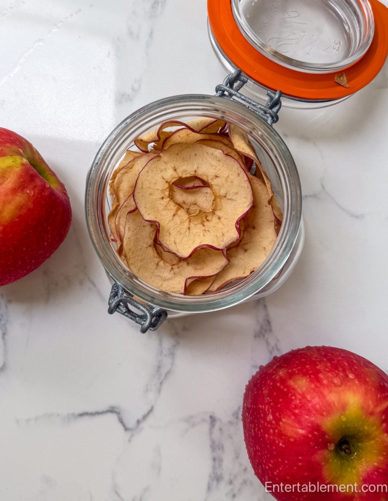 Homemade apple chips stored in a glass flip-top jar with fresh red apples on a marble countertop.