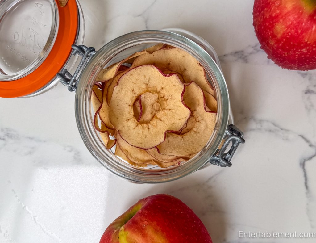 Homemade apple chips stored in a glass flip-top jar with fresh red apples on a marble countertop.