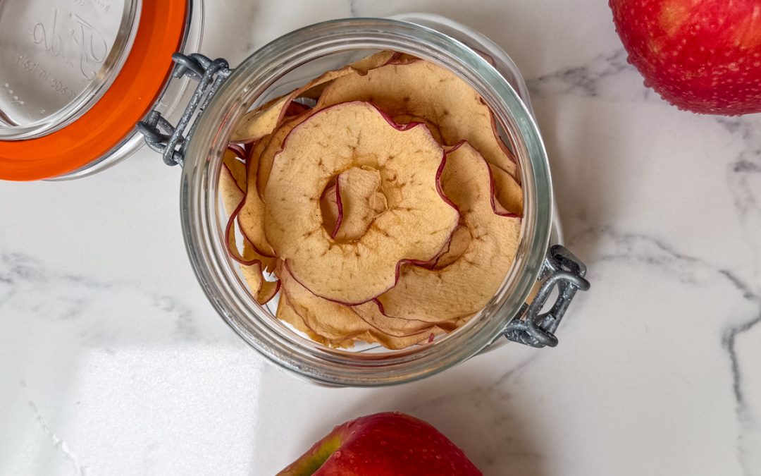 Homemade apple chips stored in a glass flip-top jar with fresh red apples on a marble countertop.