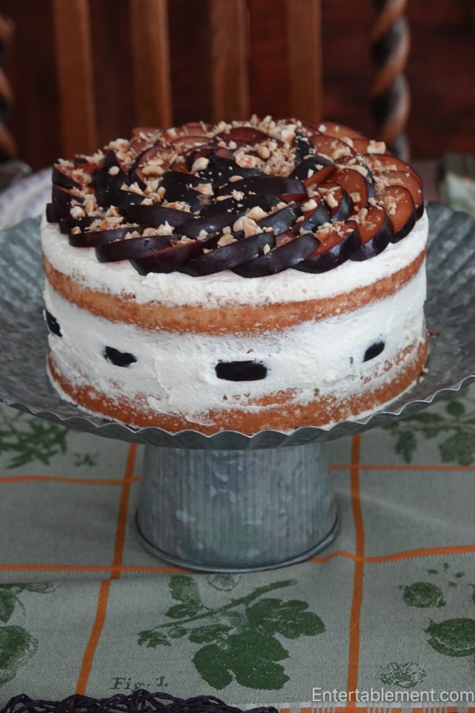 Plum Hazelnut Cake with whipped cream frosting, fresh plum slices arranged in a spiral and toasted hazelnuts, displayed on a galvanized pedestal cake stand.