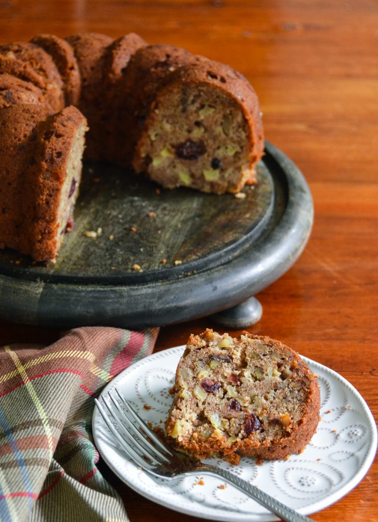 Apple, cranberry, walnut, and spice Bundt cake on a dark pedestal stand with a generous slice served on a white patterned plate.