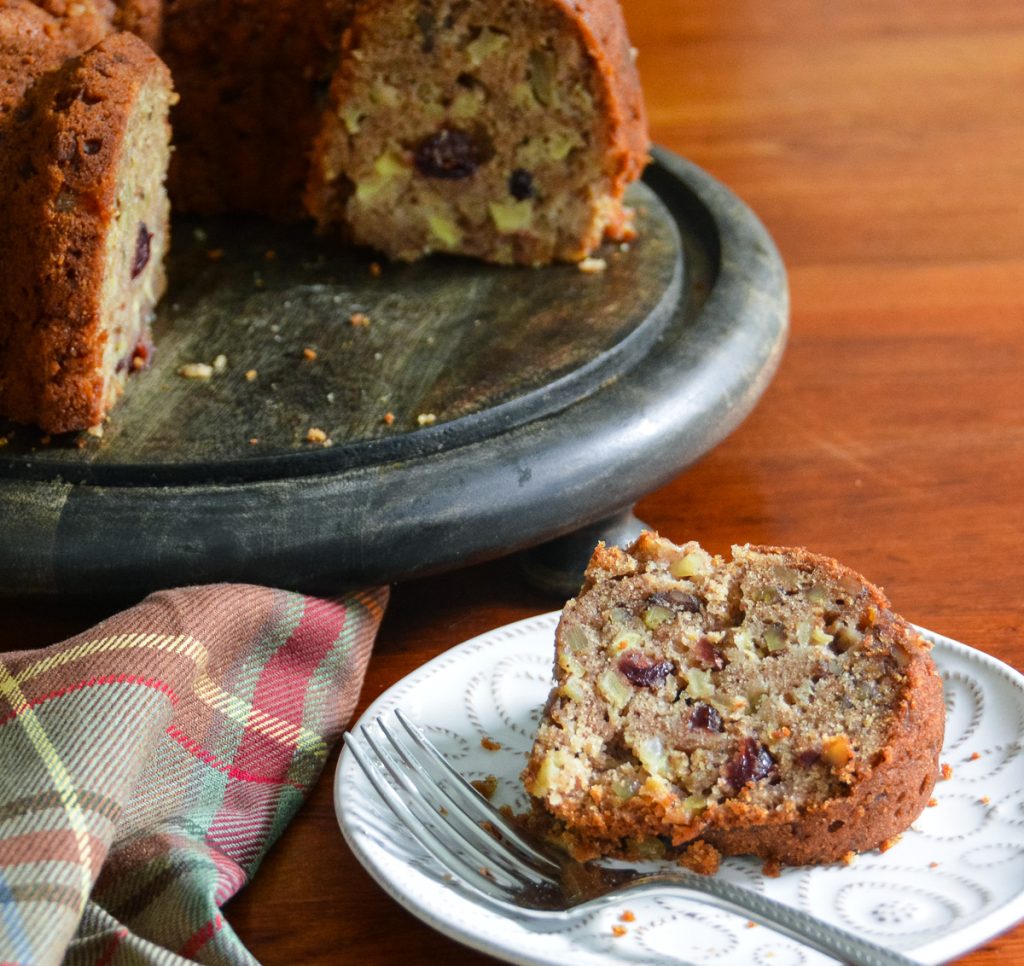 Slice of apple, cranberry, walnut, and spice Bundt cake on a white patterned plate beside a dark cake stand, with a tartan napkin and fork on a wooden table.