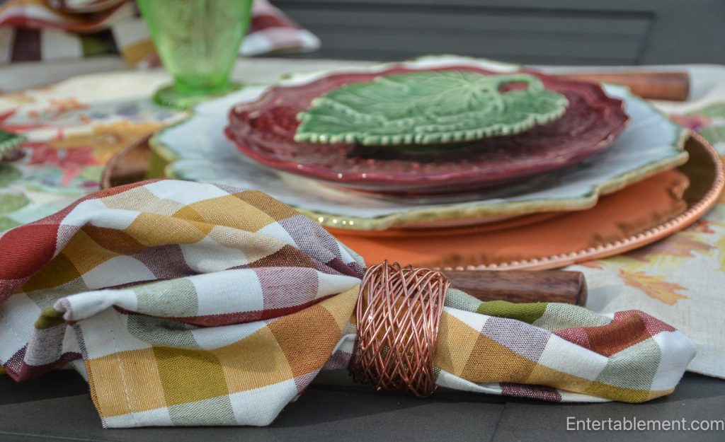Checked autumn napkin in burgundy, green and gold with an open copper napkin ring and wooden-handled flatware.