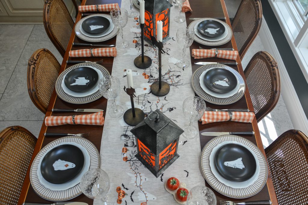 Six Halloween place settings surrounding a ghost-patterned runner with lanterns and tall candlesticks